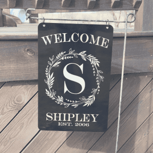 Black metal welcome sign with white engraved text and design, hanging from a metal stand. The sign reads 'WELCOME' at the top, followed by a large letter 'S' inside a decorative wreath, and the name 'SHIPLEY' with 'EST. 2006' at the bottom. The background features a wooden fence and a deck railing.