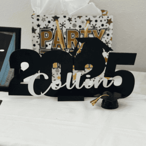 Personalized 2025 graduation name sign with the name Collin displayed on a table with a graduation cap decoration and gift bag in the background
