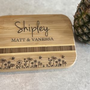A small bamboo cutting board engraved with the name "Shipley" in a handwritten-style font and "Matt & Vanessa" in an all-caps serif font. The board features a stripe of darker wood and an engraved row of wildflowers along the bottom. A fresh pineapple sits to the right on a light gray marble countertop.