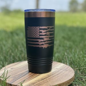 Black 20 oz tumbler with engraved copper American flag made of rifles, displayed outdoors on a wooden base with grass background