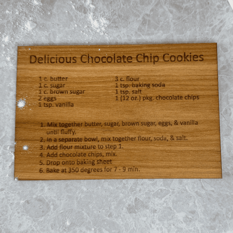 Wooden recipe card engraved with “Delicious Chocolate Chip Cookies” ingredients and instructions, lying on a floured marble countertop.