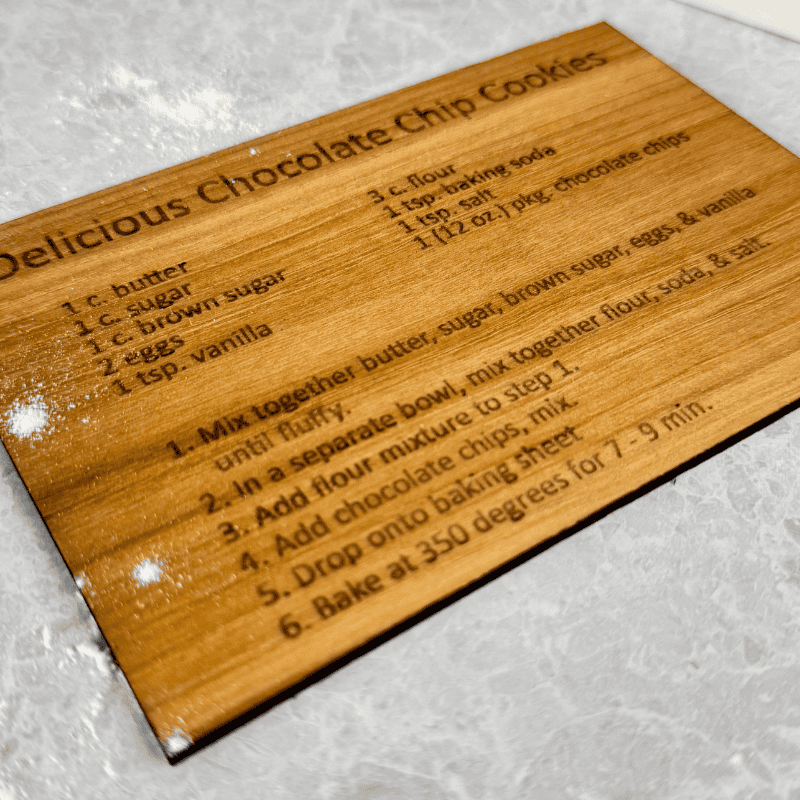 Laser-engraved wooden recipe card showing clear cookie recipe text on a light wood surface with flour dusting.