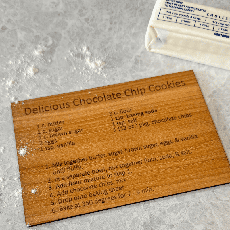 Personalized wooden recipe card with engraved chocolate chip cookie recipe, shown at an angle on a marble surface with flour and butter in view.