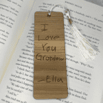 Wooden bookmark engraved with child’s handwriting that reads “I love you Grandma – Ella,” finished with a white tassel, resting on an open book.