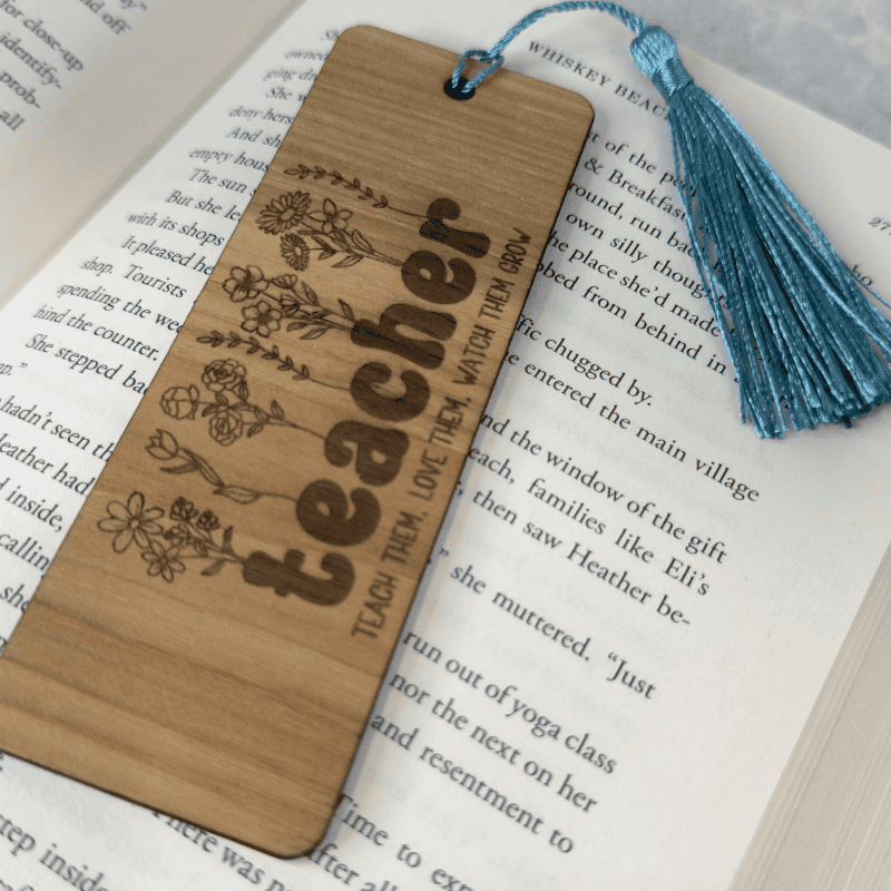 Wooden teacher bookmark engraved with floral design and the phrase “Teach them. Love them. Watch them grow,” finished with a blue tassel on an open book.