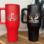 Red and black insulated tumblers engraved with antlers and lettering in silver laser etching, displayed on a wooden tabletop.