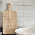 engraved handwritten potato salad recipe on a wooden cutting board displayed against a white subway tile kitchen backsplash